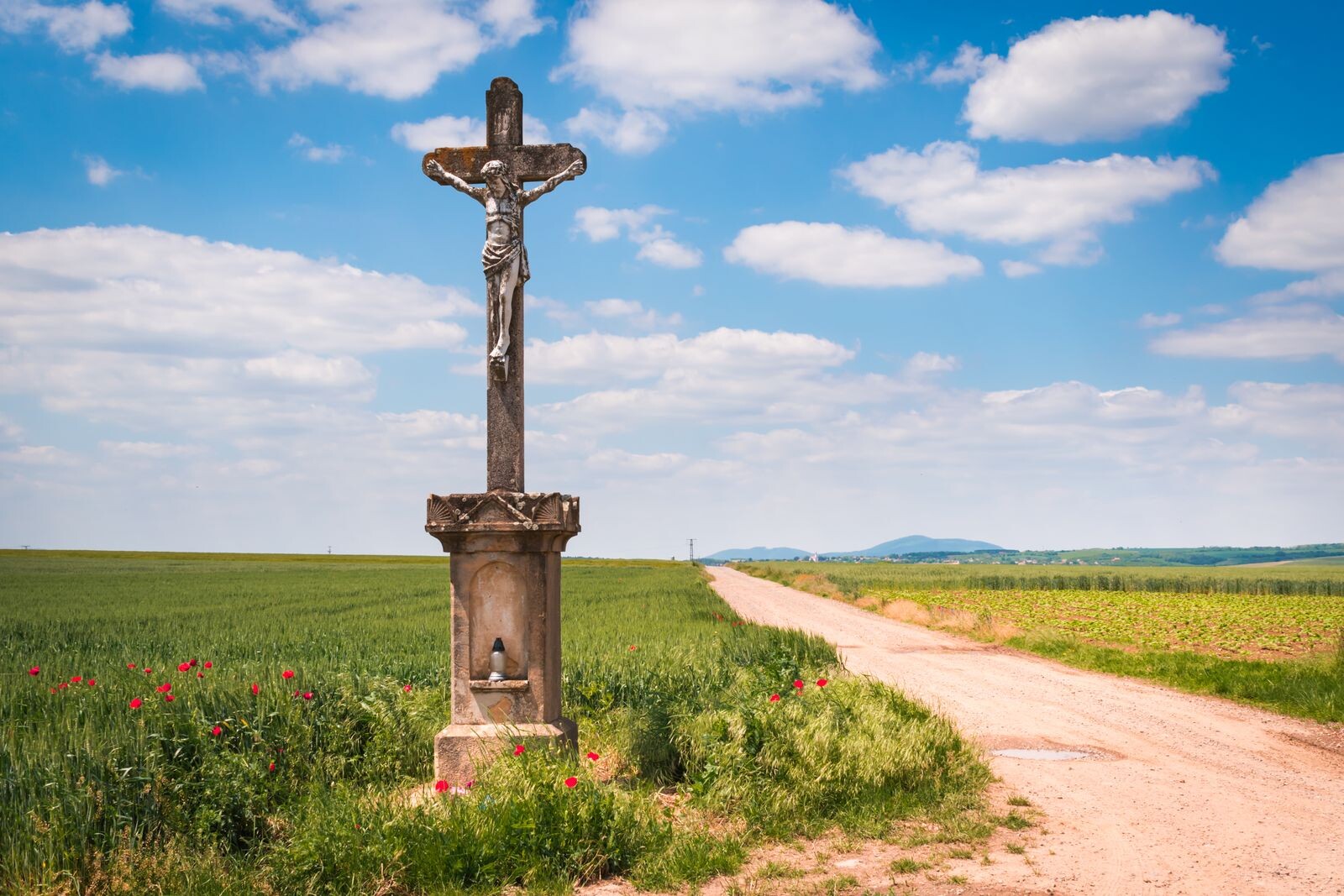 A serene rural scene with a crucifix at a crossroads, fields, and a blue sky.
